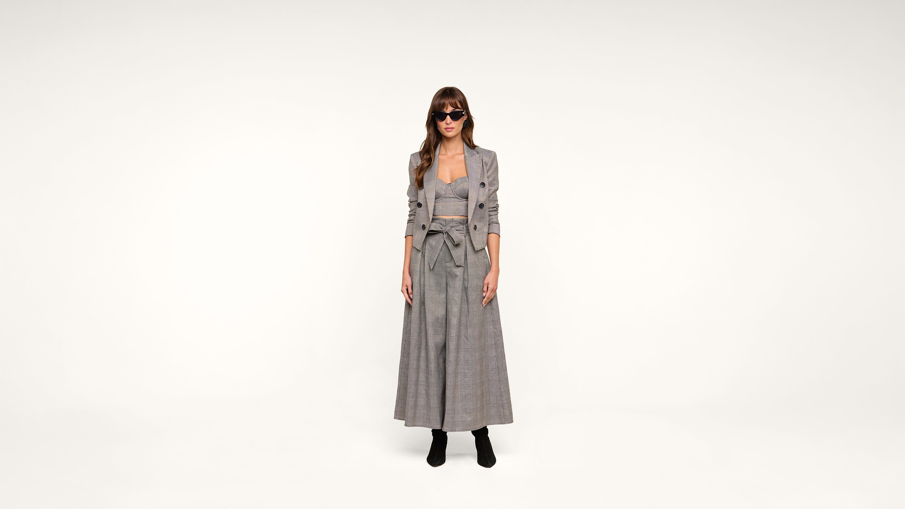 A woman wearing sunglasses, a long plaid dress, and a matching blazer stands against a plain white background. She has long hair and is wearing black ankle boots.
