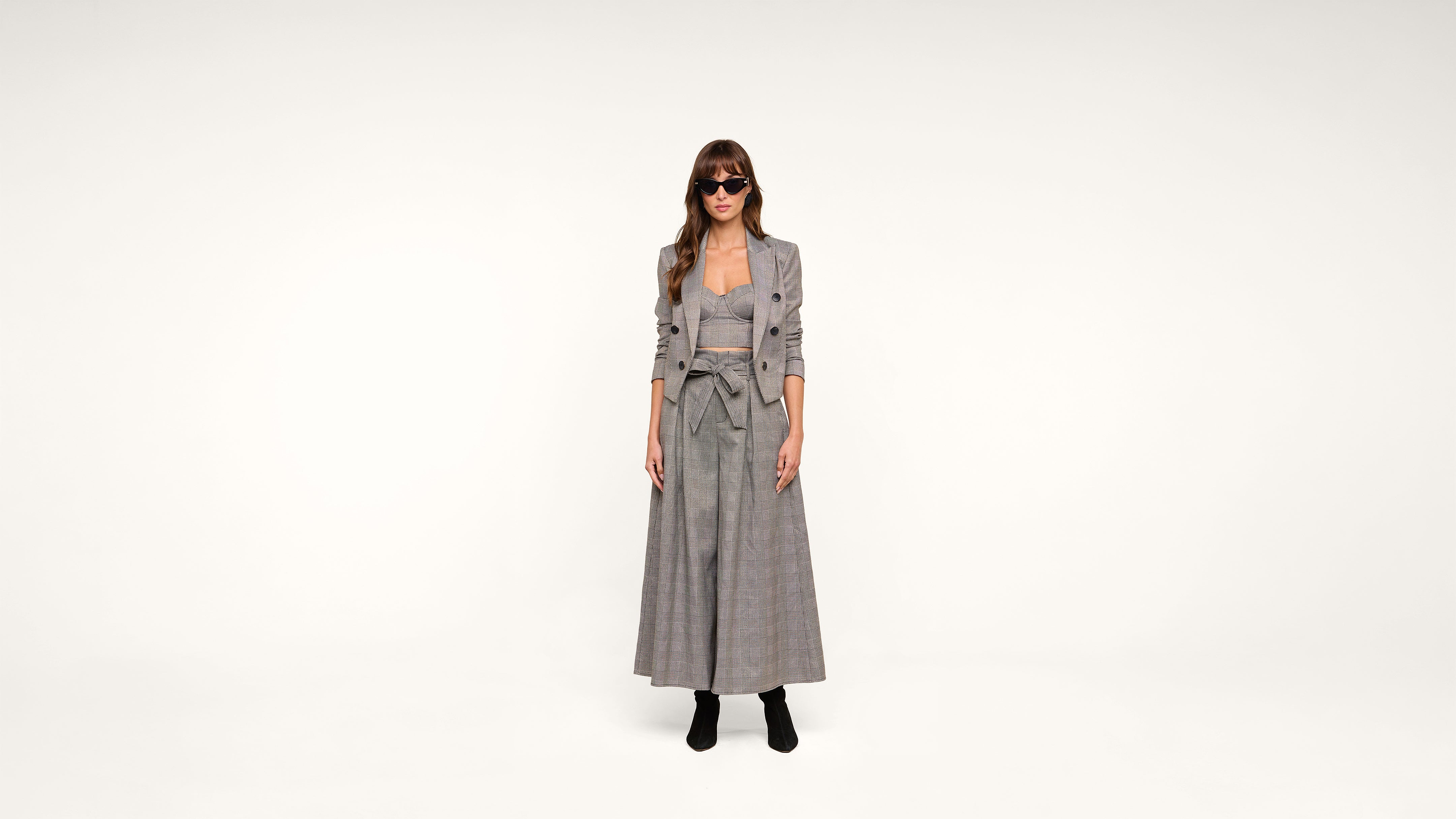 A woman wearing sunglasses, a long plaid dress, and a matching blazer stands against a plain white background. She has long hair and is wearing black ankle boots.