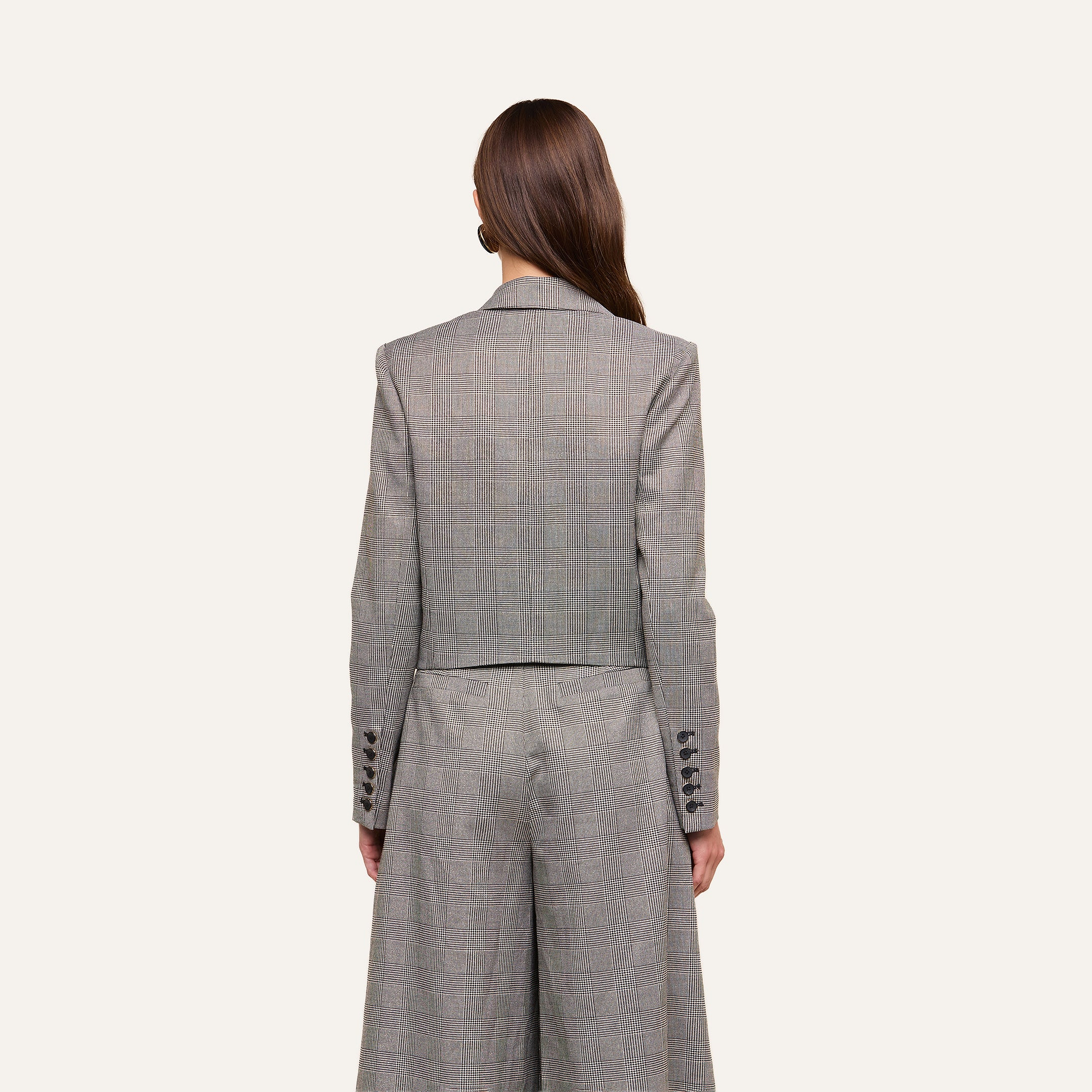 A woman with long brown hair, seen from behind, wears the Lila Tweed Blazer in a gray plaid pattern paired with matching wide-leg trousers. The background is plain and light-colored.