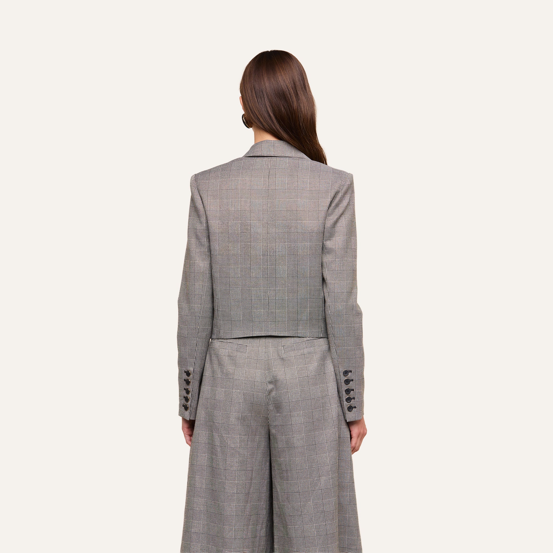 A woman with long brown hair, seen from behind, wears the Lila Tweed Blazer in a gray plaid pattern paired with matching wide-leg trousers. The background is plain and light-colored.