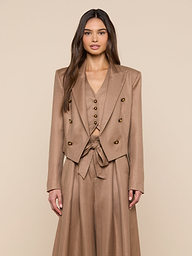 A woman with long brown hair wears the Lila Linen-Blend Blazer, part of a stylish tan three-piece suit with a double-breasted blazer, buttoned vest, and high-waisted skirt with tied belt, standing against a beige background.