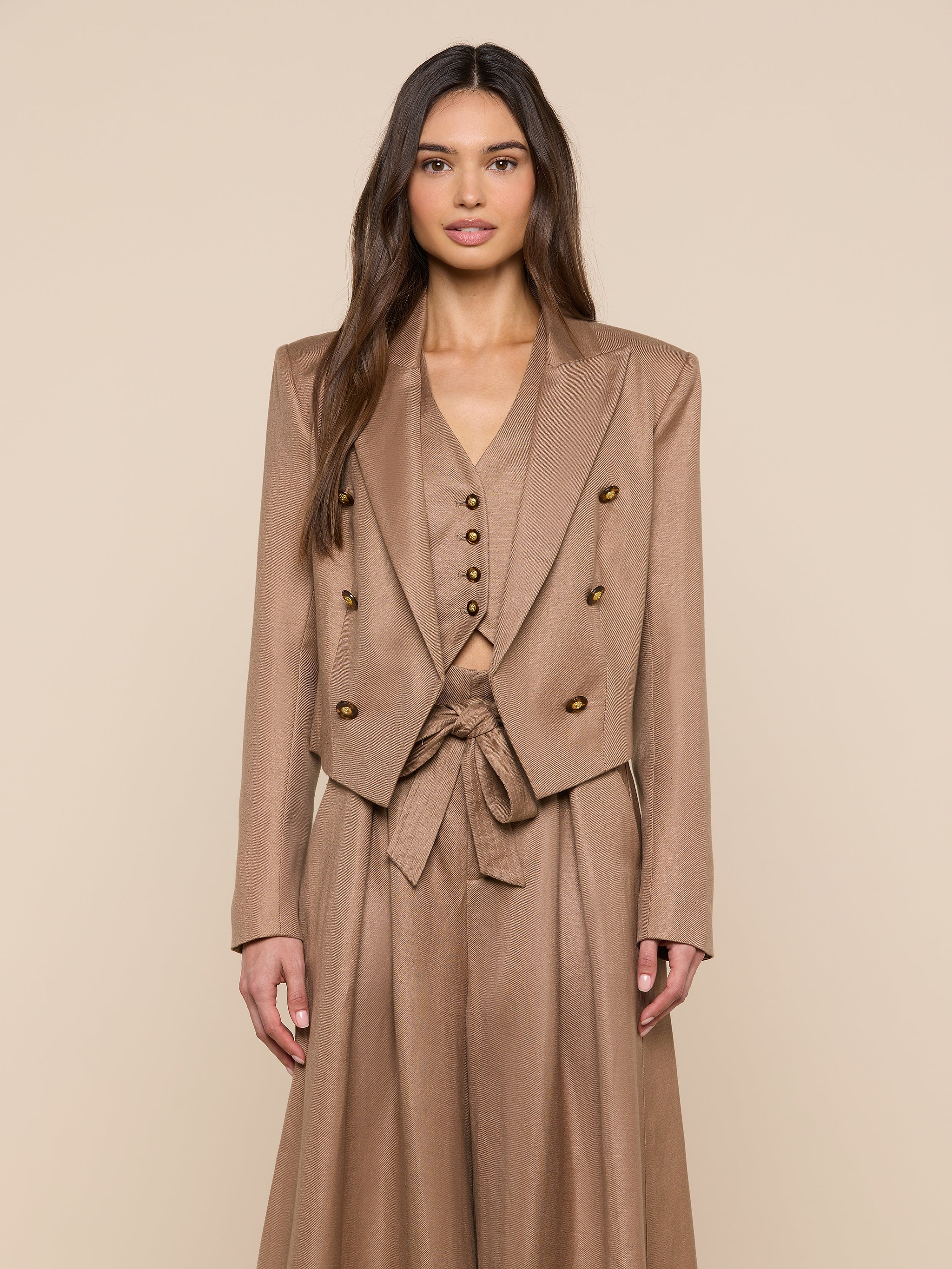 A woman with long brown hair wears the Lila Linen-Blend Blazer, part of a stylish tan three-piece suit with a double-breasted blazer, buttoned vest, and high-waisted skirt with tied belt, standing against a beige background.