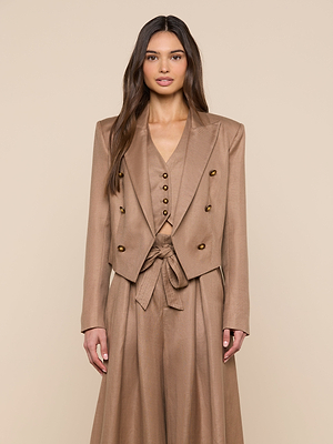 A woman with long brown hair wears the Lila Linen-Blend Blazer, part of a stylish tan three-piece suit with a double-breasted blazer, buttoned vest, and high-waisted skirt with tied belt, standing against a beige background.