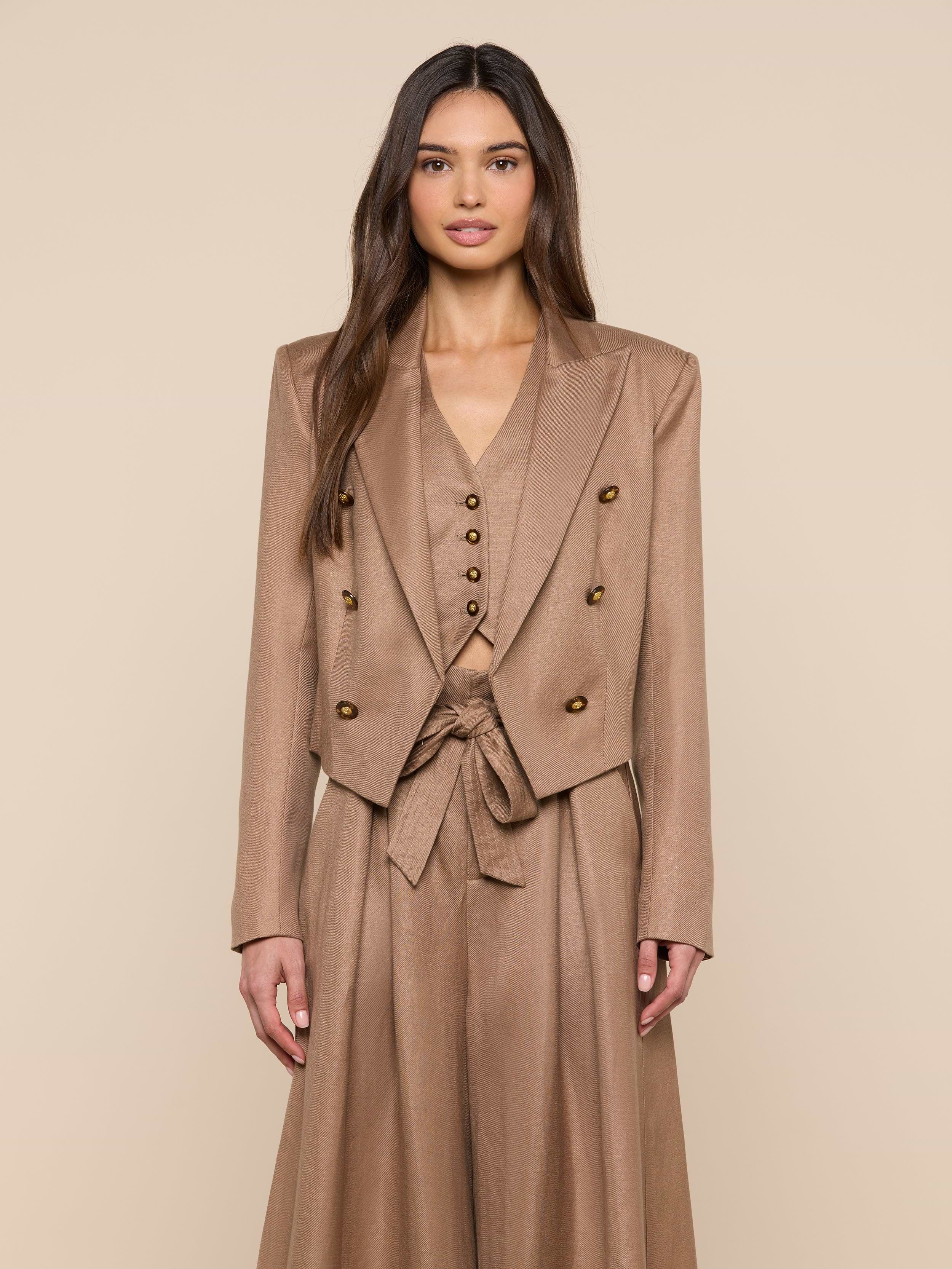 A woman with long brown hair wears the Lila Linen-Blend Blazer, part of a stylish tan three-piece suit with a double-breasted blazer, buttoned vest, and high-waisted skirt with tied belt, standing against a beige background.