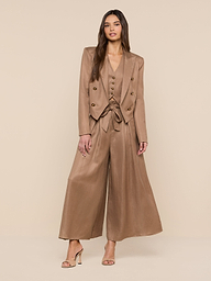 A woman with long hair poses confidently against a beige background, wearing the Lila Linen-Blend Blazer styled as a modern pantsuit with a belted waist, wide-leg trousers, and high-heeled sandals.