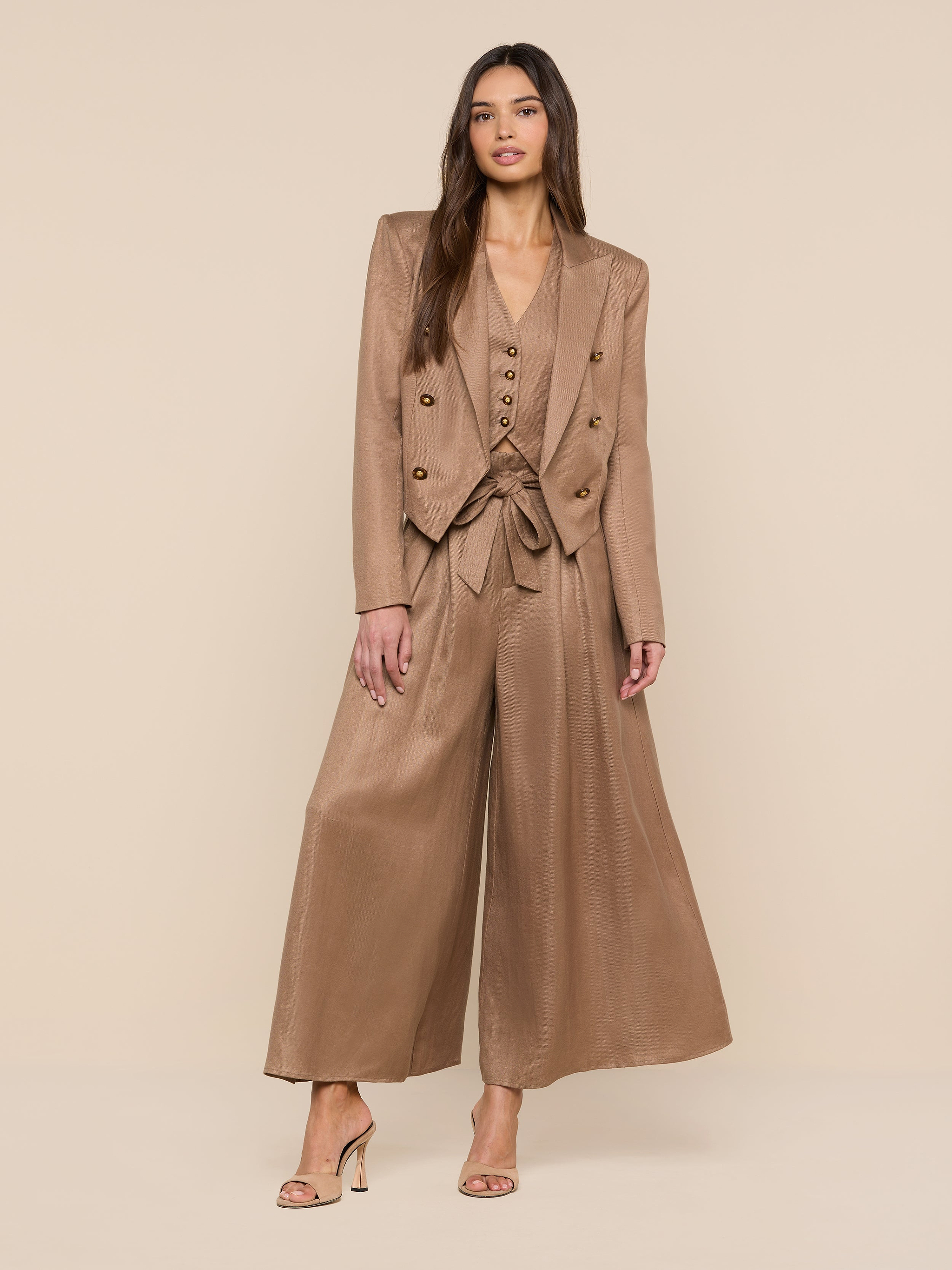 A woman with long hair poses confidently against a beige background, wearing the Lila Linen-Blend Blazer styled as a modern pantsuit with a belted waist, wide-leg trousers, and high-heeled sandals.