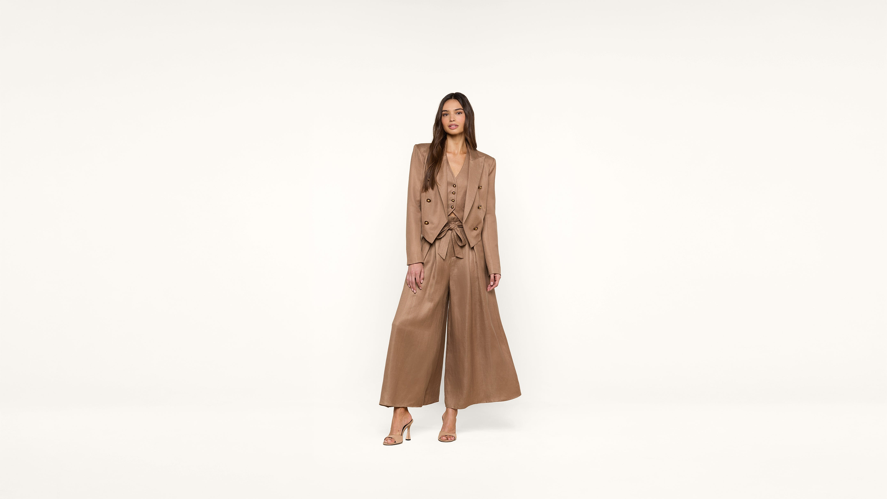 A woman stands against a plain white background wearing a tan suit with wide-leg pants, a matching vest, high heels, and a blazer. She has long brown hair and a neutral expression.