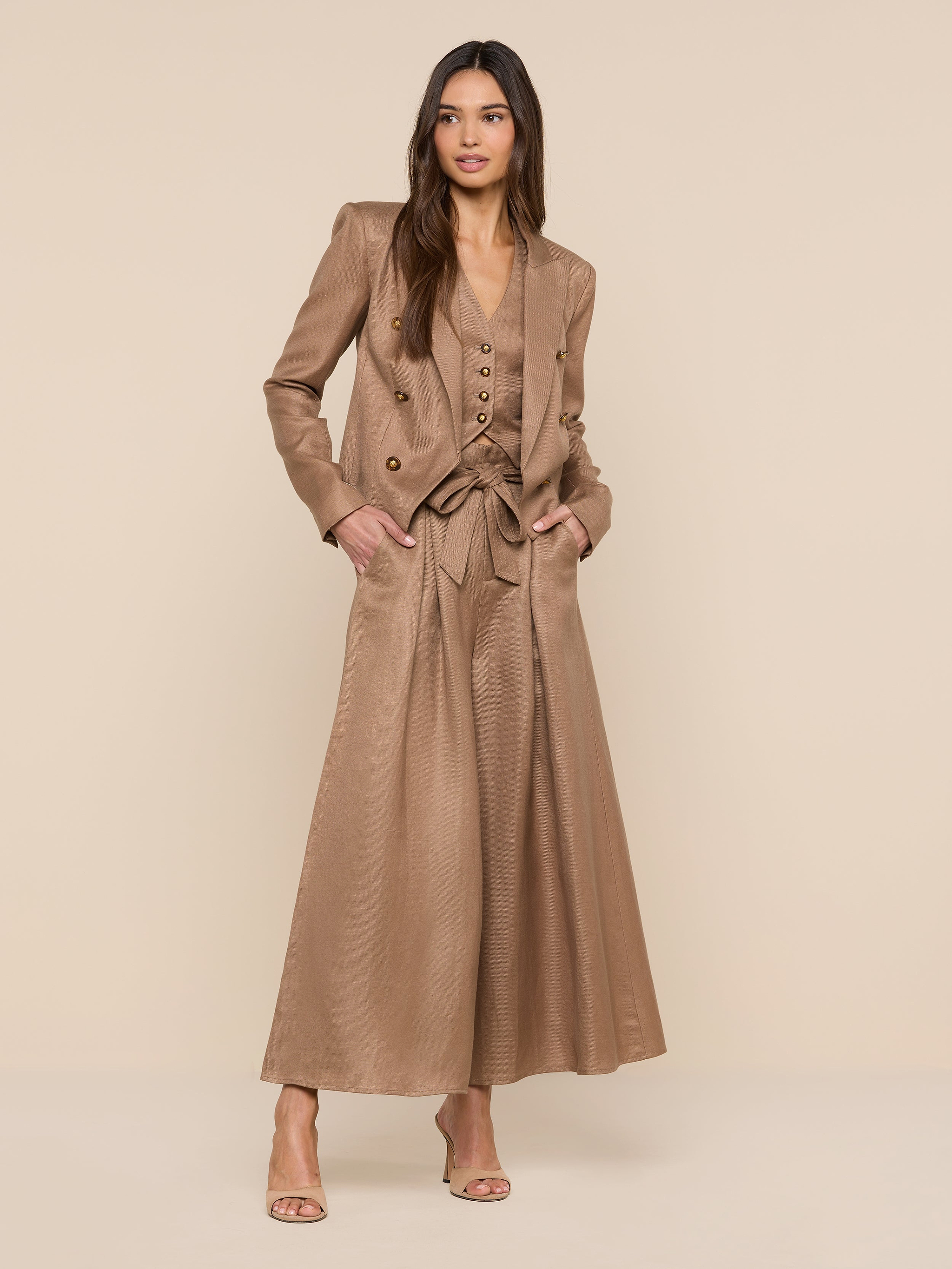 A woman stands against a beige background wearing the Lila Linen-Blend Blazer as part of a light brown, wide-leg pantsuit with a belted vest. She has long hair, hands in her pockets, and open-toed heels.