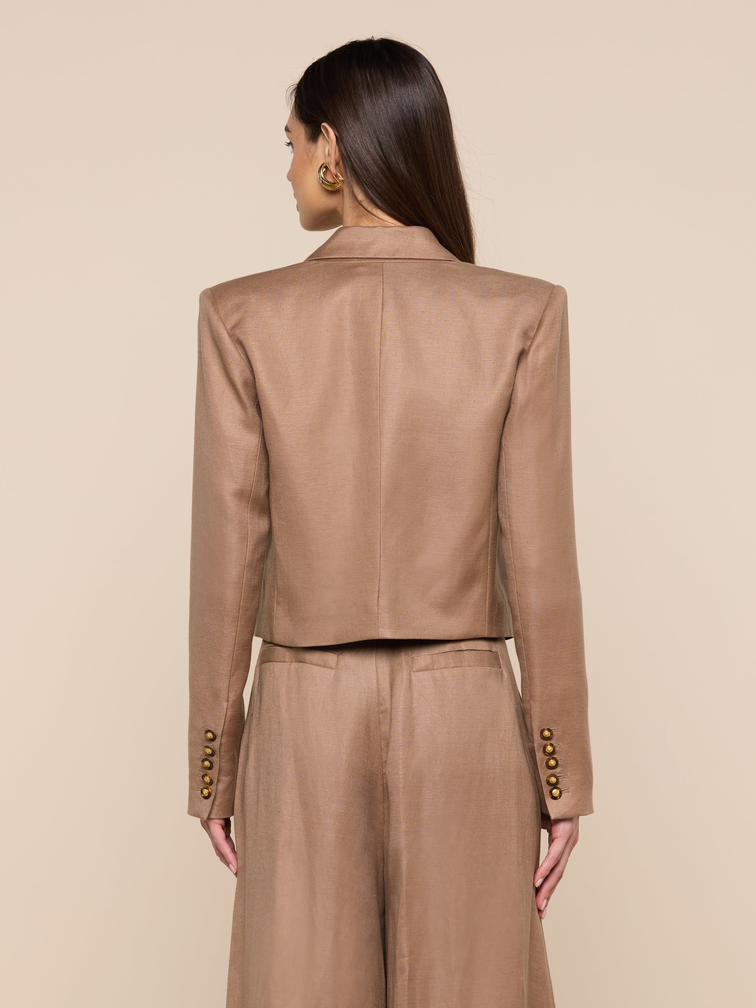 A woman with long brown hair stands facing away, wearing the Lila Linen-Blend Blazer and matching brown trousers. The smooth texture and gold cuff buttons create an elegant look against the beige background.
