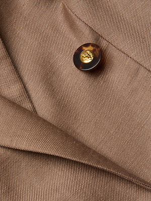 Close-up of the Lila Linen-Blend Blazer in brown, showcasing a tortoiseshell button with a gold center and overlapping fabric folds featuring a visible diagonal texture.