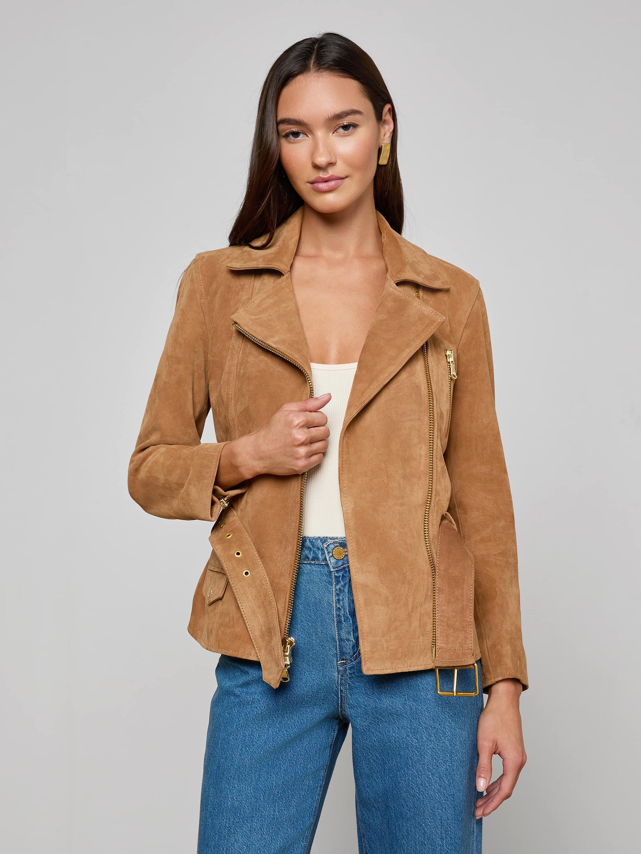 A woman wearing the Dinah Suede Biker Jacket in tan over a white top and blue jeans stands against a plain light background, holding one side of the women’s outerwear open.