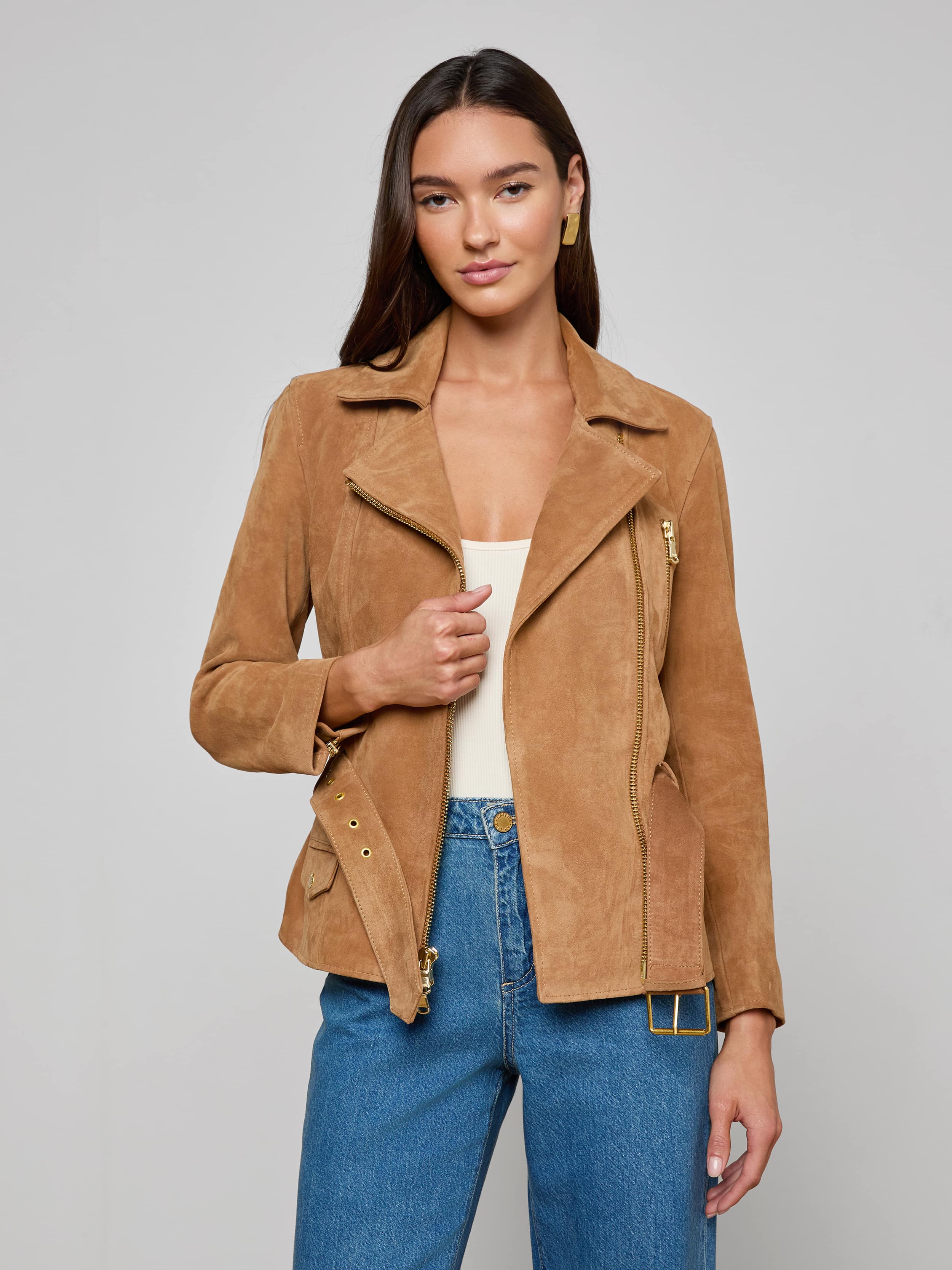 A woman wearing the Dinah Suede Biker Jacket in tan over a white top and blue jeans stands against a plain light background, holding one side of the women’s outerwear open.