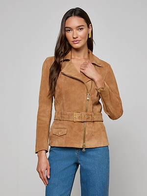 A woman with long brown hair and a neutral expression stands against a plain background wearing the Dinah Suede Biker Jacket, featuring gold zips, an adjustable belt, and blue jeans.