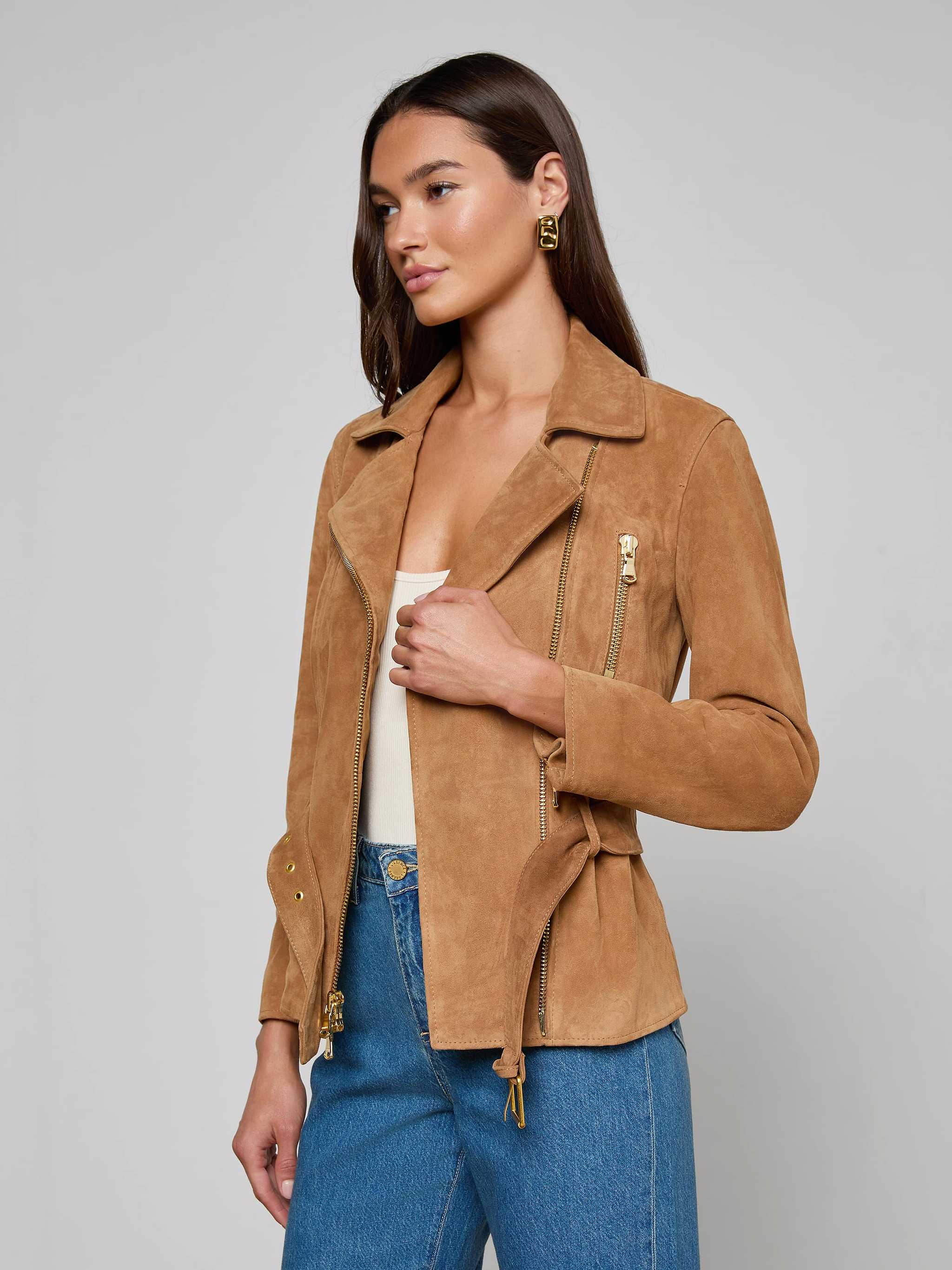 A woman stands against a plain light background, looking to the side with one hand holding her Dinah Suede Biker Jacket, worn over a white top and blue jeans.
