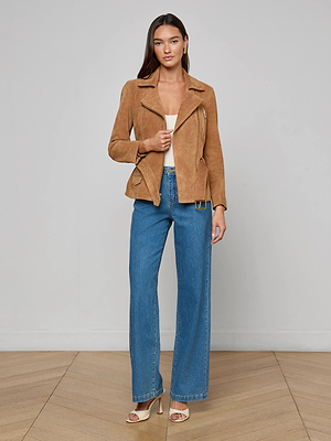 A woman with long brown hair models the Dinah Suede Biker Jacket in tan with gold zips, styled over a white top, blue wide-leg jeans, and beige heeled sandals, standing on a wooden floor against a light gray wall.