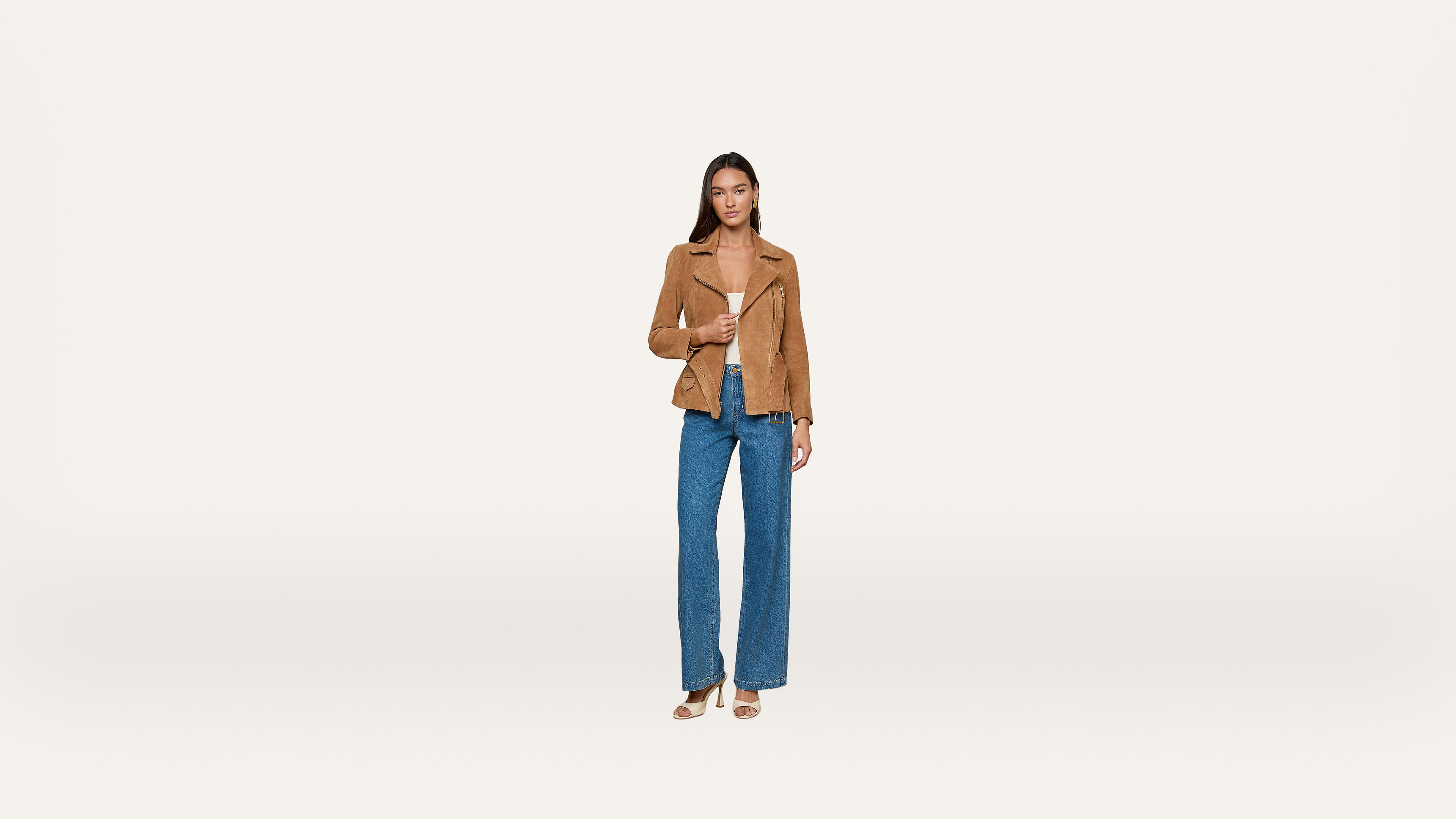 A woman stands facing forward, wearing a brown jacket, blue wide-leg jeans, and heeled sandals against a plain white background.