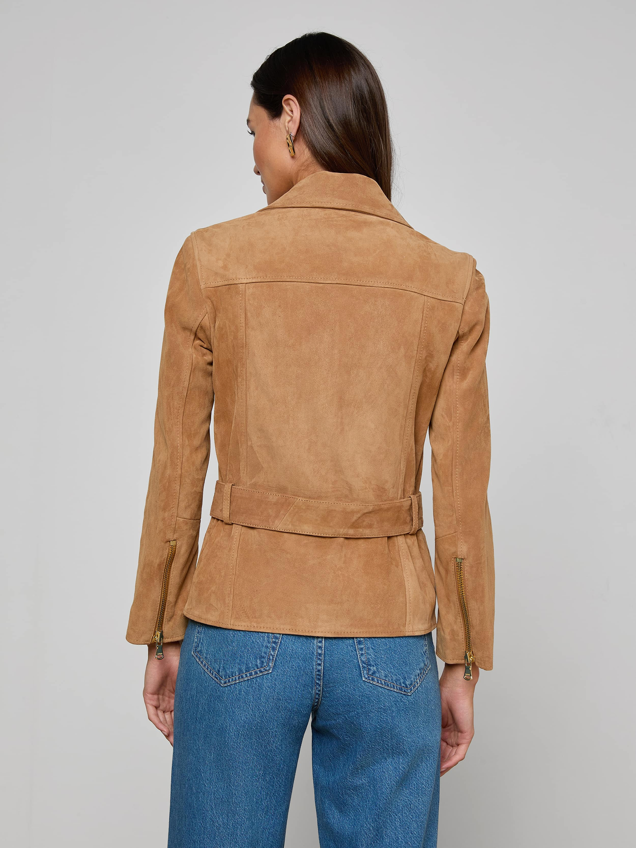 A woman with long brown hair is seen from behind, wearing the Dinah Suede Biker Jacket—a tan suede belted jacket—paired with blue jeans. She stands against a plain light background, showcasing the stylish women’s outerwear.