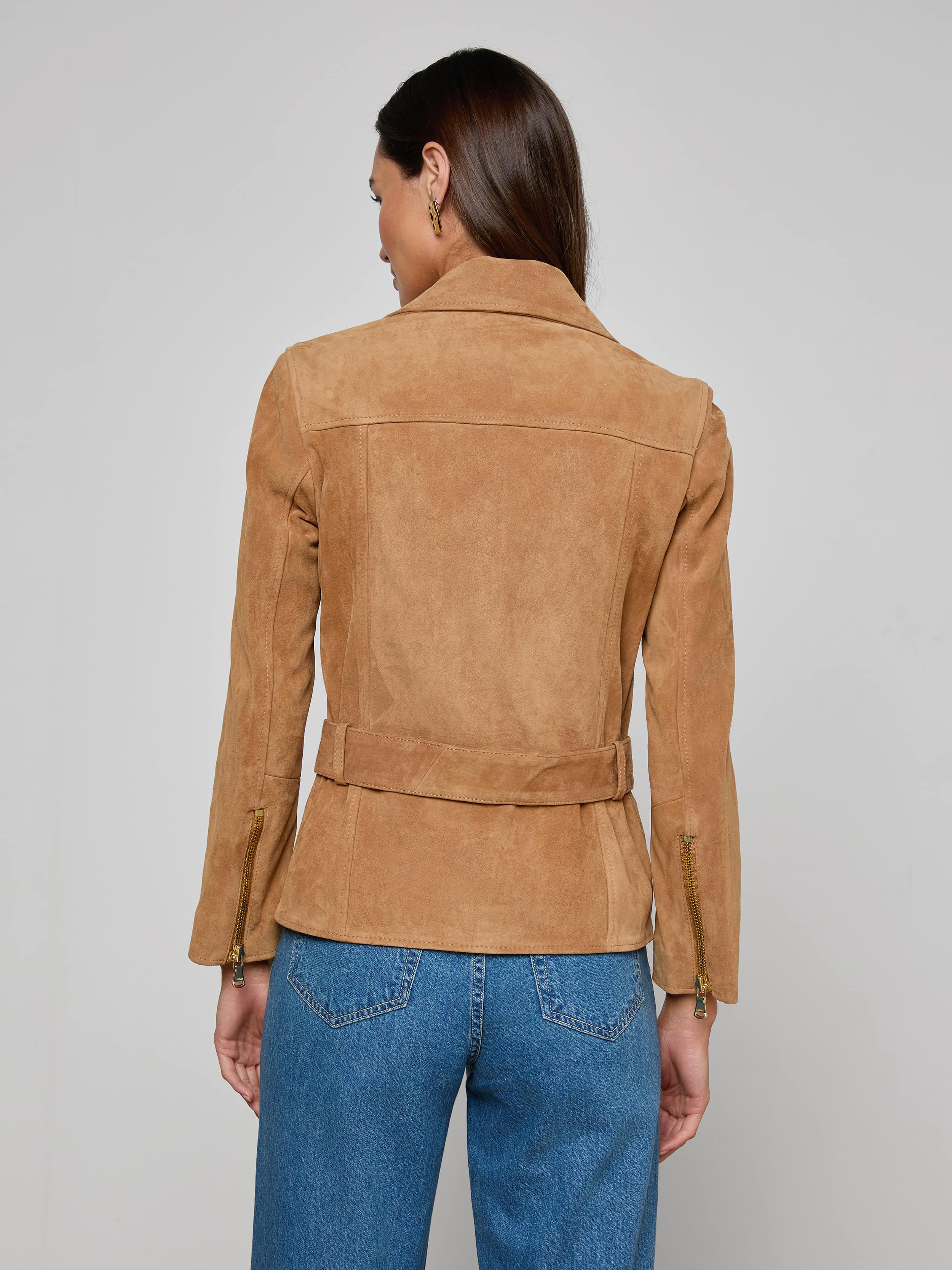 A woman with long brown hair is seen from behind, wearing the Dinah Suede Biker Jacket—a tan suede belted jacket—paired with blue jeans. She stands against a plain light background, showcasing the stylish women’s outerwear.