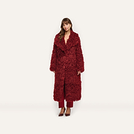 A woman wears a long, deep red Lizbeth Faux Fur Jacket over matching trousers and pointed-toe heels.
