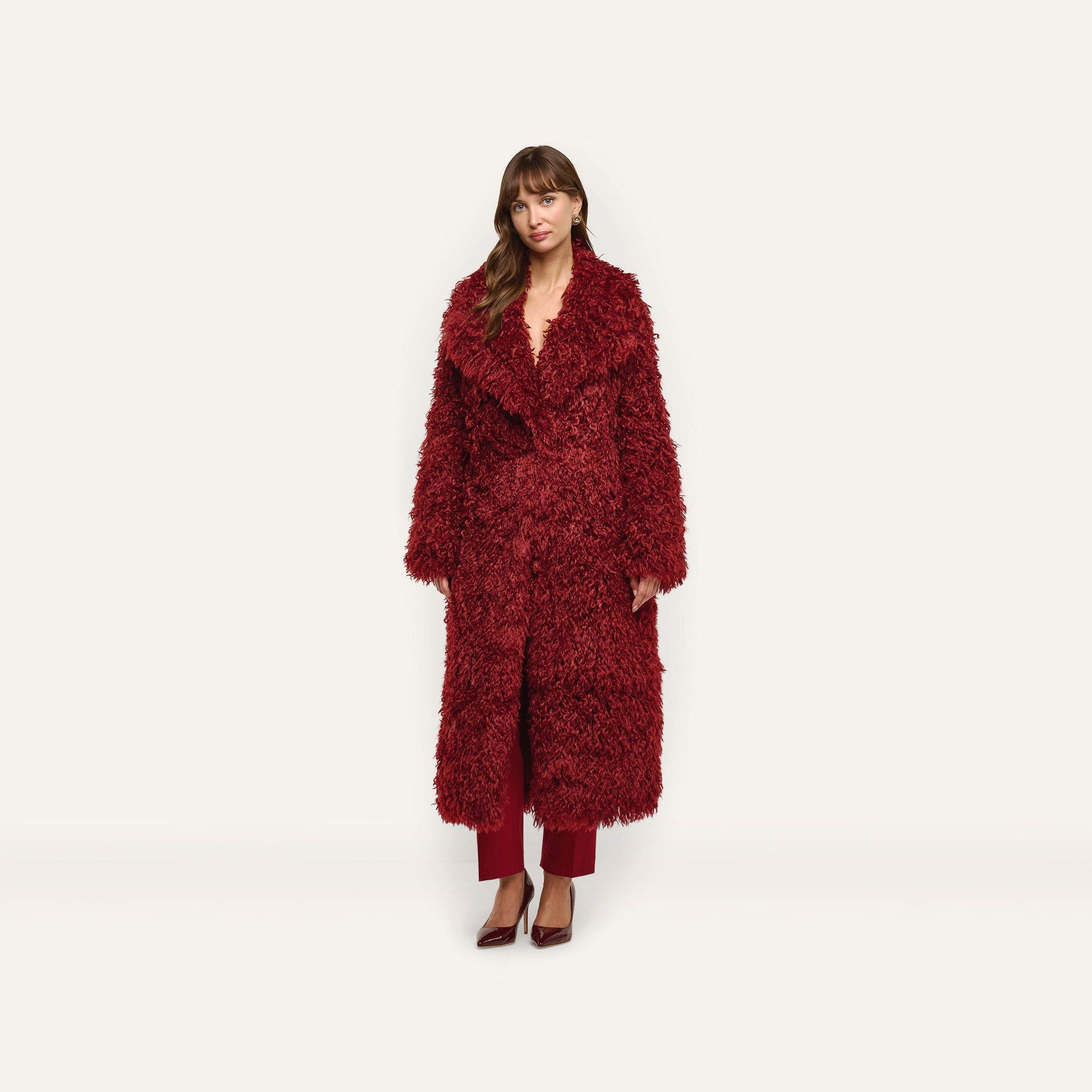 A woman wears a long, deep red Lizbeth Faux Fur Jacket over matching trousers and pointed-toe heels.