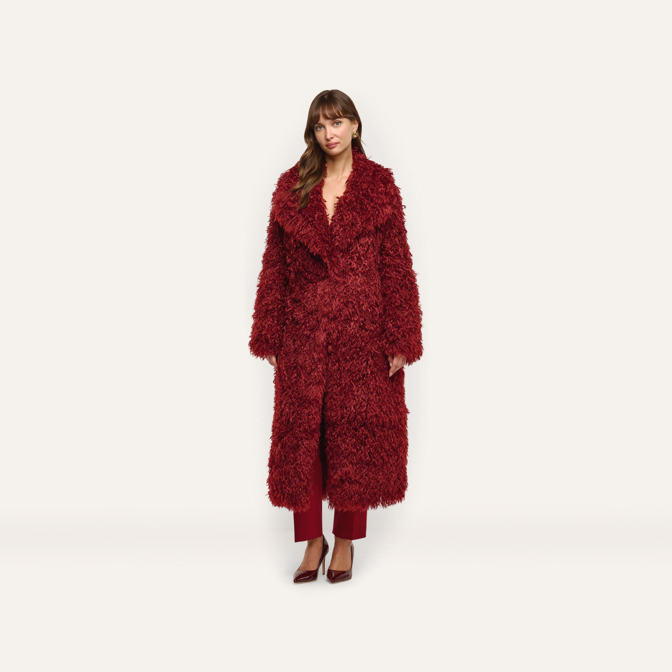 A woman wears a long, deep red Lizbeth Faux Fur Jacket over matching trousers and pointed-toe heels.