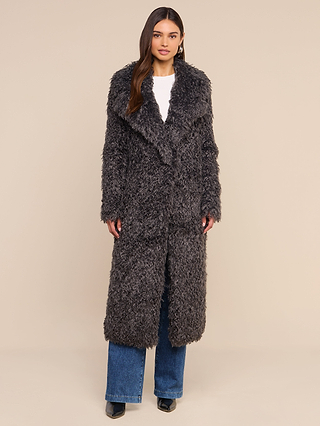 A woman stands facing forward against a plain beige background, wearing the Lizbeth Faux Fur Jacket in a long, curly-textured dark gray shag over a white top and blue jeans, paired with black shoes.