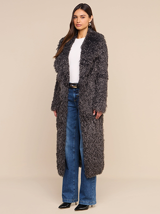 A woman stands against a plain beige background wearing the Lizbeth Faux Fur Jacket—a long, textured gray shag coat—over a white top and blue jeans with black boots, embodying the effortless cool of New York Fashion Week.