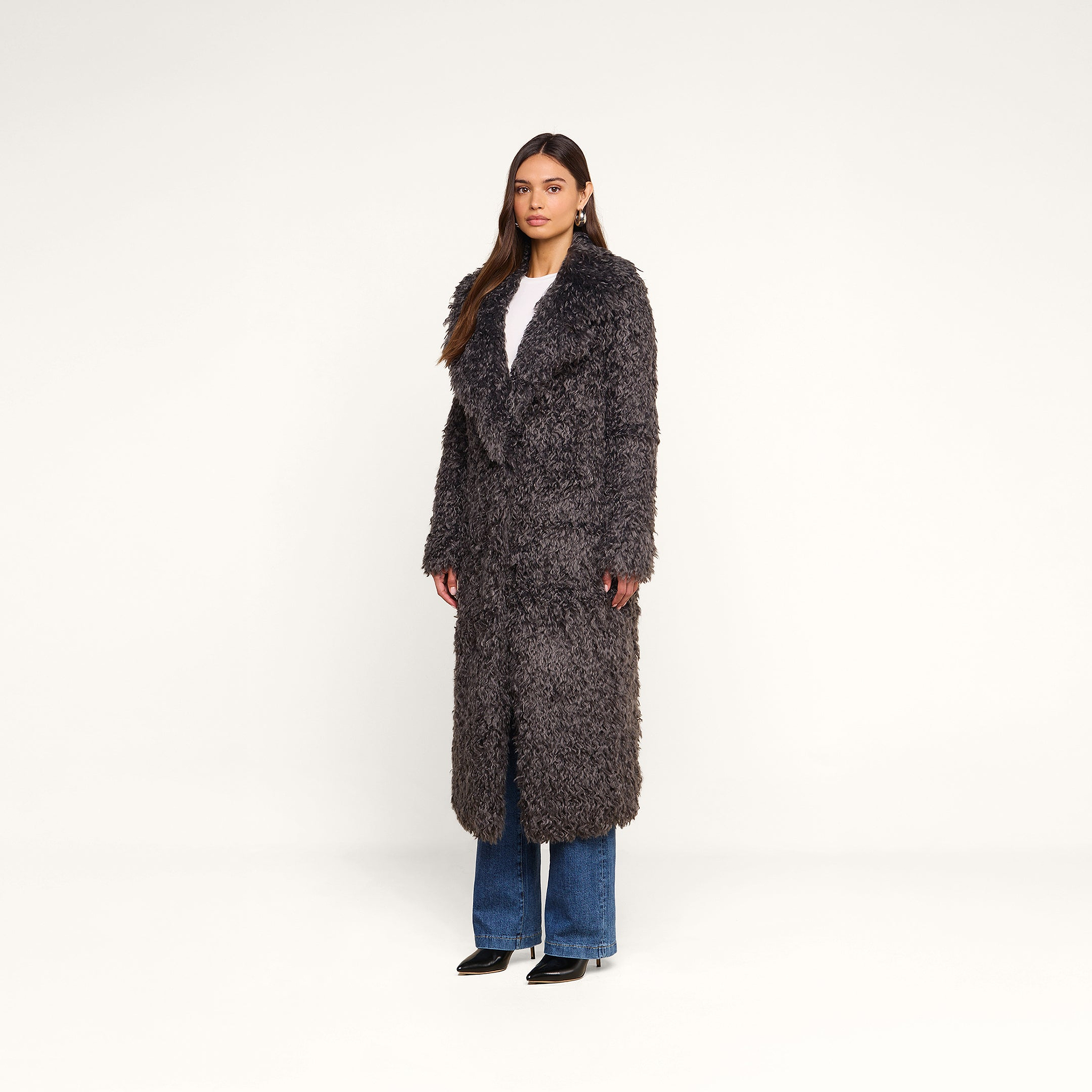 A woman stands against a plain white background wearing the Lizbeth Faux Fur Jacket in gray over a white top and blue jeans with black shoes.