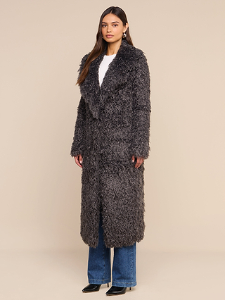 A woman stands against a plain beige background wearing the Lizbeth Faux Fur Jacket—a long, textured, dark gray shag faux fur coat—layered over a white top, blue jeans, and black pointed shoes.