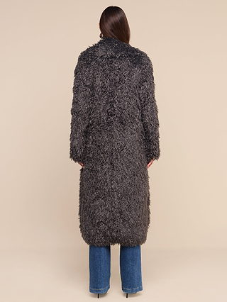 A person with long brown hair wears the Lizbeth Faux Fur Jacket in a textured dark gray shag, styled in a relaxed fit with blue jeans. They stand with their back to the camera against a plain beige background, capturing an understated look reminiscent of New York Fashion Week street style.