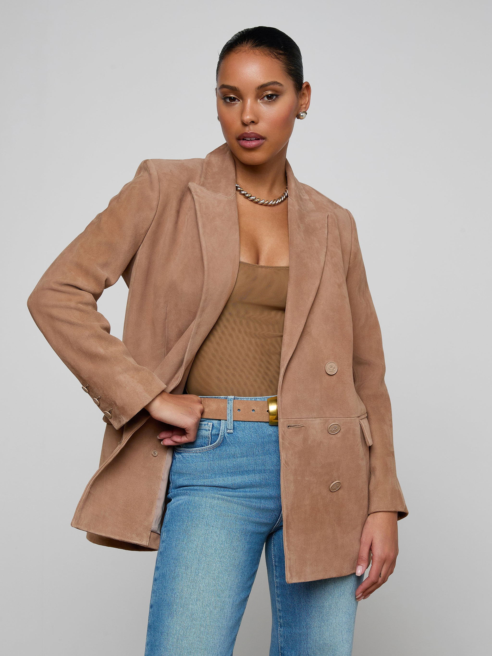 Wearing the Aimee Oversized Suede Blazer in tan over a brown top and blue jeans, accessorized with a gold necklace, a woman stands against a plain light background, gazing confidently at the camera.