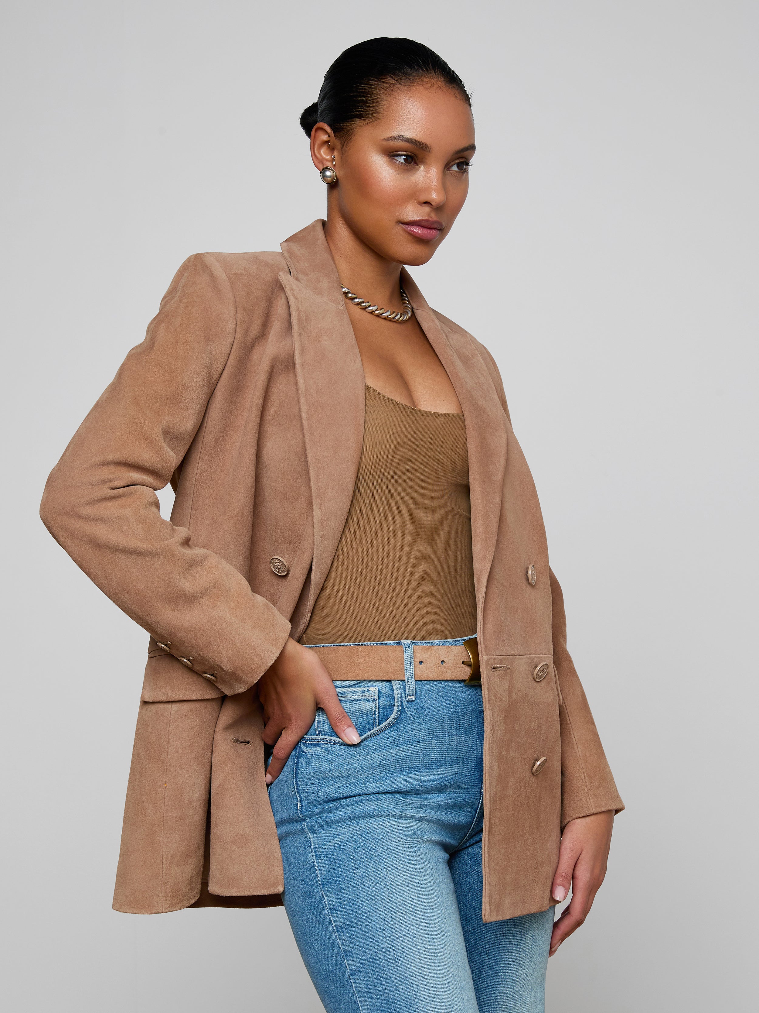 A woman poses with one hand on her hip and looks to the side against a plain light background, styled in the Aimee Oversized Suede Blazer layered over a brown top and light blue jeans.