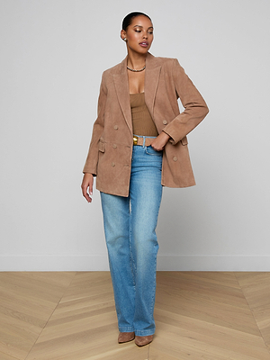 A woman with short hair poses against a plain wall, wearing the Aimee Oversized Suede Blazer over a brown top and blue jeans, accessorized with earrings, a necklace, and brown heels, one hand in her pocket.