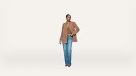 A person stands against a plain white background, wearing a light brown blazer, tan top, blue jeans, and brown shoes, with one hand in their pocket and the other relaxed by their side.