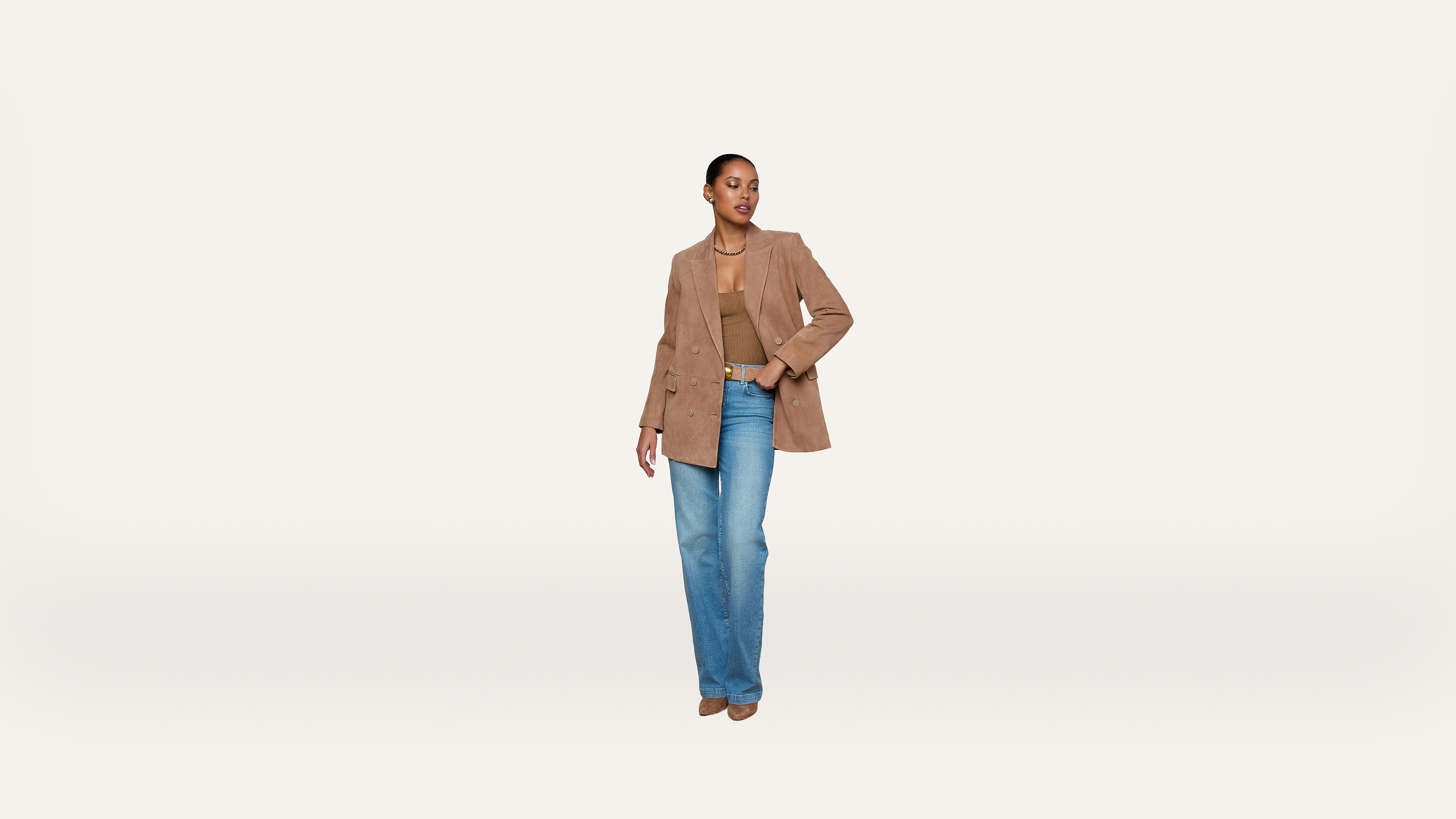 A person stands against a plain white background, wearing a light brown blazer, tan top, blue jeans, and brown shoes, with one hand in their pocket and the other relaxed by their side.
