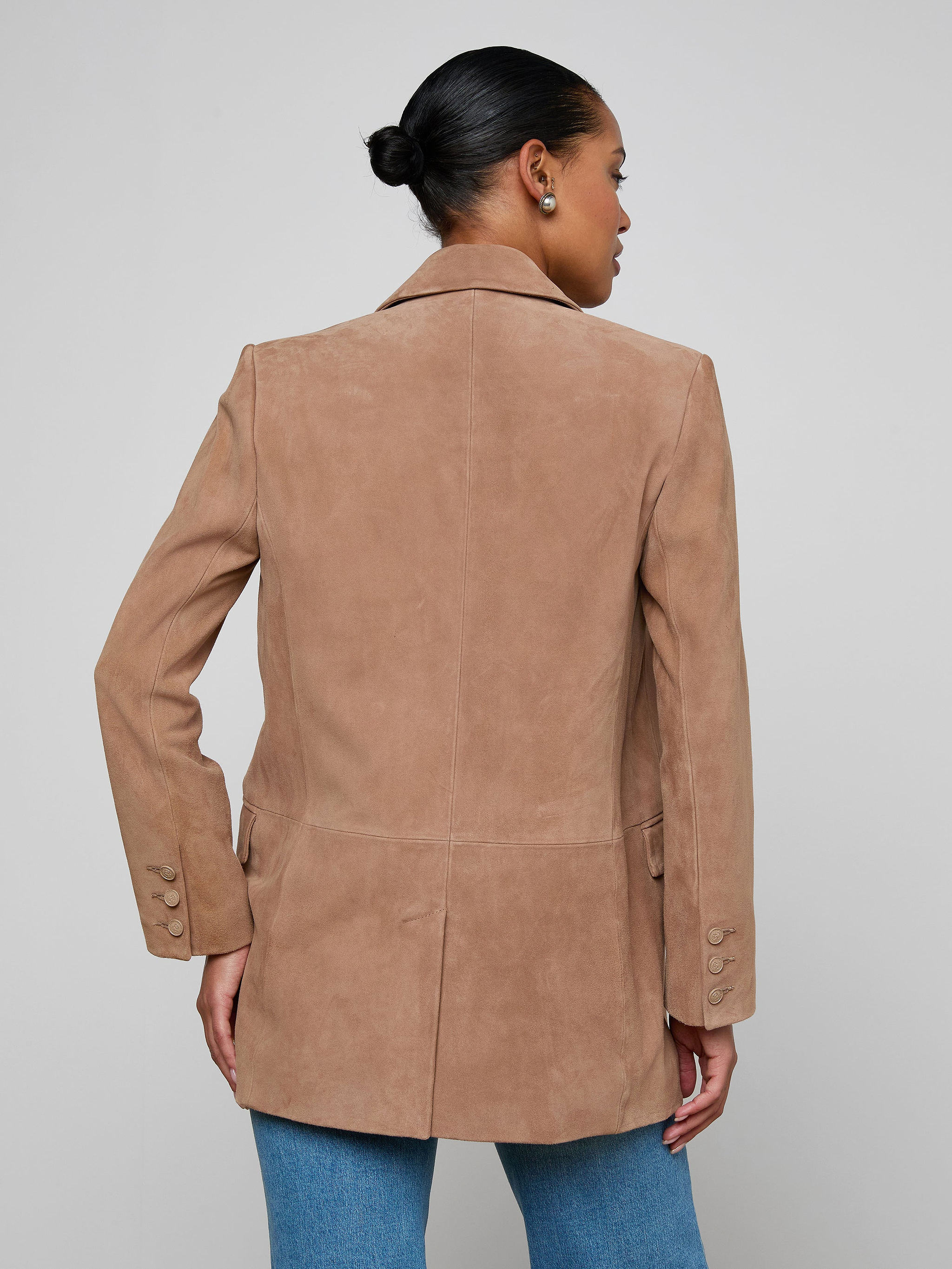 A woman with her hair in a bun wears the Aimee Oversized Suede Blazer in tan and blue jeans, facing away from the camera against a plain light background.