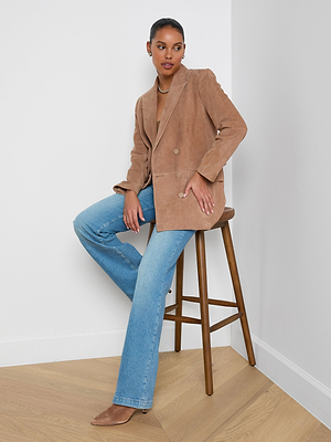 A woman with short hair sits on a wooden stool against a white wall, wearing the Aimee Oversized Suede Blazer over a matching top, light blue jeans, and tan pointed-toe shoes. She accessorizes with a necklace.
