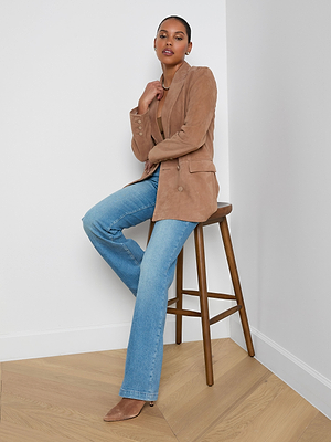 A woman sits on a wooden stool wearing the Aimee Oversized Suede Blazer, paired with a light brown top, blue jeans, and tan heels. She poses against a white wall and wood floor, with one hand near her chin and the other on her knee.