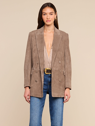A woman stands against a plain beige background wearing the Aimee Oversized Suede Blazer in taupe over a beige blouse with a deep neckline, paired with blue jeans and a black belt featuring a gold buckle.