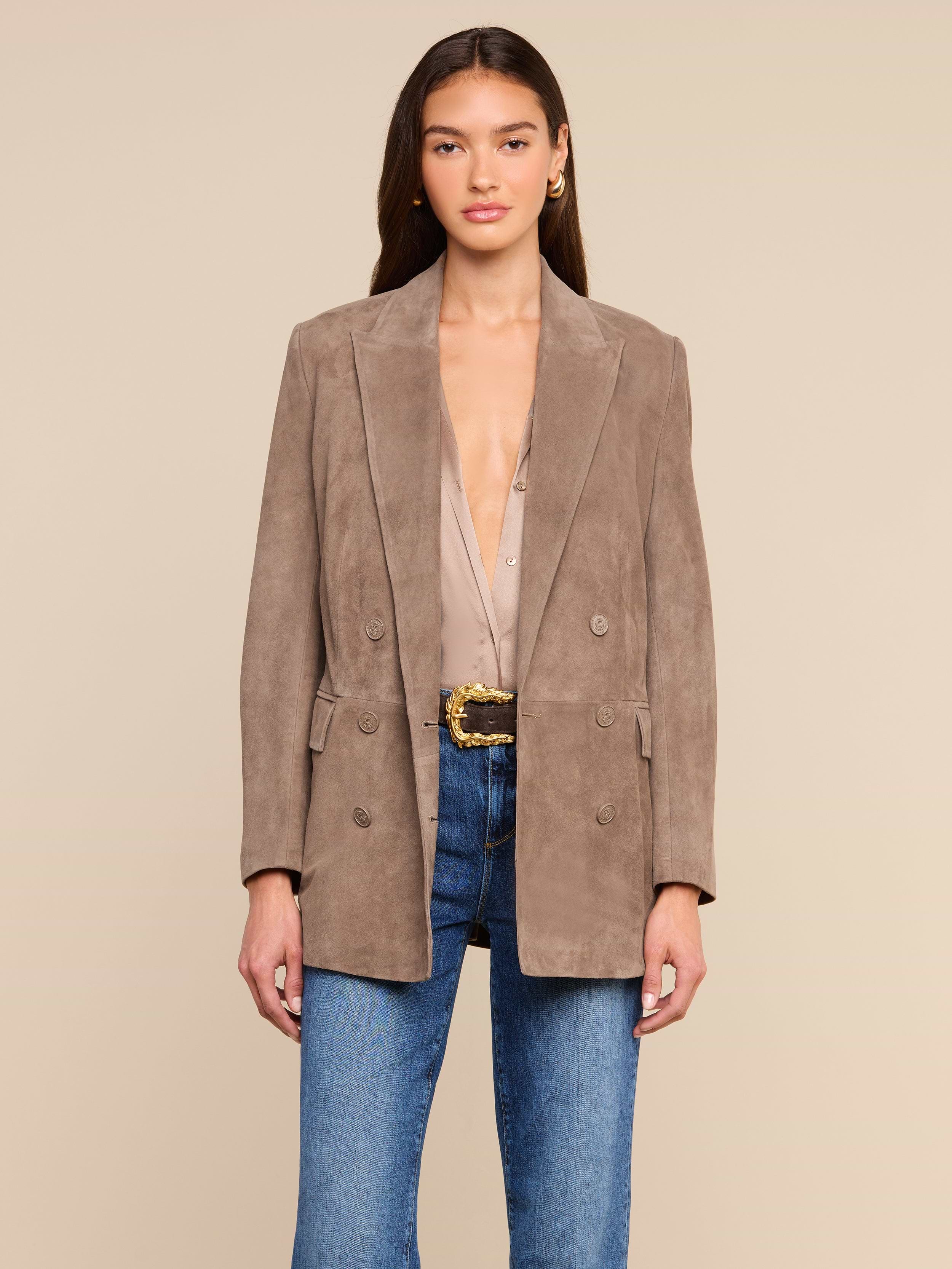 A woman wearing the Aimee Oversized Suede Blazer in taupe over a beige blouse, paired with blue jeans and a textured belt, stands against a plain light background, looking forward with her hands at her sides.