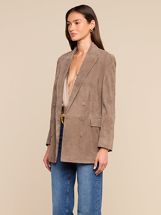 A woman wearing the Aimee Oversized Suede Blazer in taupe, paired with a light beige deep-neck top, blue jeans, and gold hoop earrings stands against a plain beige background.