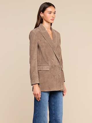 A woman stands against a beige background, wearing the Aimee Oversized Suede Blazer, paired with gold earrings and blue jeans. She faces slightly to the right with a neutral expression.