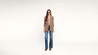 A woman stands against a white background wearing sunglasses, a tan blazer, a low-cut top, blue jeans, and brown heels.