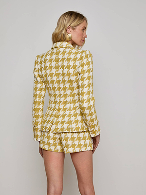 A woman with blonde hair and gold hoop earrings stands facing away, wearing the Marie Houndstooth Tweed Blazer in yellow and white with matching shorts against a plain light background.