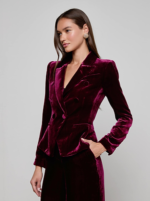 A woman with long brown hair models the Marie Velvet Blazer by L'AGENCE in maroon, paired with matching pants. She stands against a plain light background, facing slightly sideways with one hand at her side.