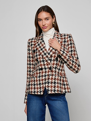 A woman with long brown hair stands confidently against a plain background, wearing the Marie Houndstooth Tweed Blazer over a white turtleneck and blue jeans.