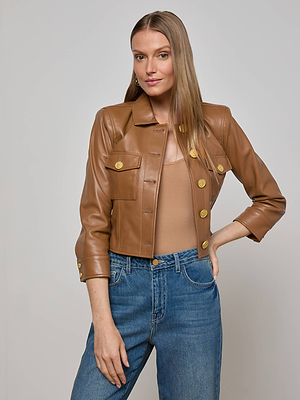A woman with long, straight hair models the L'AGENCE Kumi Cropped Leather Jacket over a beige top and blue jeans, standing against a plain light gray background with one hand in her pocket and a slight smile.