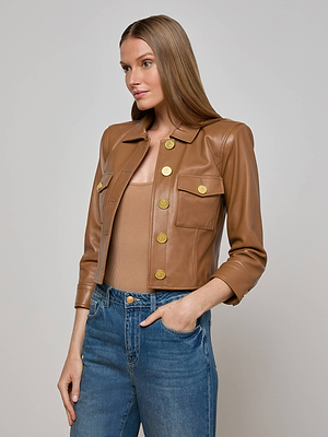 A woman with long straight hair wears the L'AGENCE Kumi Cropped Leather Jacket in brown with gold buttons over a tan top and blue jeans, standing against a plain light gray background.