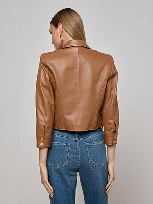 A woman with straight blond hair tucked behind her ear and gold hoop earrings wears the L'AGENCE Kumi Cropped Leather Jacket in tan faux leather with gold sleeve buttons and blue jeans, standing against a plain light gray background.