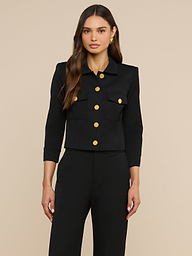 Eine Frau mit langen, welligen braunen Haaren trägt die „Kumi Cropped Jacket“ – eine schwarze, taillierte Jacke mit Dreiviertelärmeln, goldenen Knöpfen und Pattentaschen –, dazu eine passende schwarze Hose, und steht vor einem schlichten beigen Hintergrund.